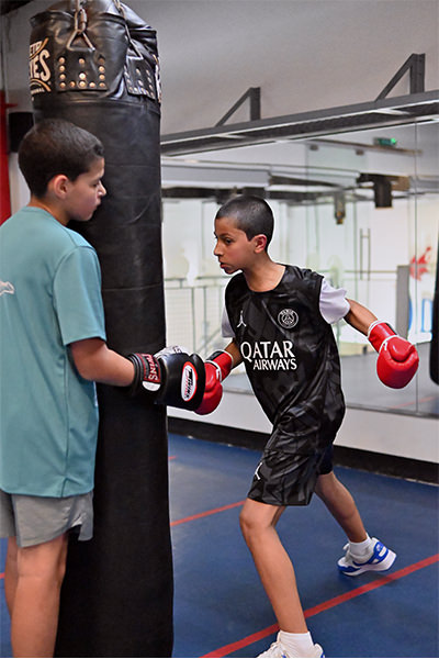 Within the hallowed halls of our esteemed boxing sanctuary in Dubai, two young pugilists hone their skills with unwavering dedication. Clad in distinctive red gloves, one adeptly delivers powerful blows to the robust heavy bag, unleashing a flurry of calculated strikes. Meanwhile, his steadfast comrade stands resolute, providing essential stability to refine their technique with precision and mutual respect. Together, they embody the spirit of discipline and perseverance that defines our gym's legacy.