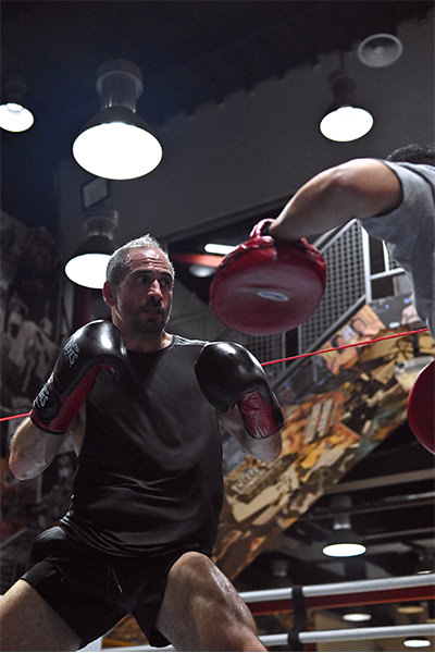 Within the elite confines of Dubai's premier boxing sanctuary, a dedicated fighter clad in sleek black attire perfects his striking prowess. Guided by his skilled partner wielding vivid red mitts, each punch is executed with precision and power. Overhead, stylish hanging lights complement the gym's vibrant mural backdrop, symbolizing the dynamic energy and world-class expertise that define our establishment.