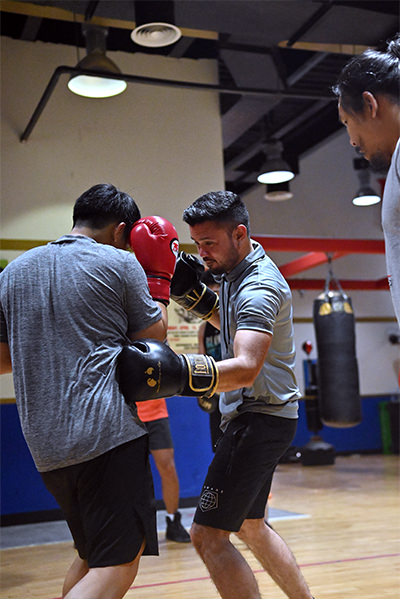 Within the elite confines of our distinguished Dubai boxing academy, two athletes gracefully engage in a dynamic sparring session. One commands attention with red gloves, while their formidable counterpart competes adorned in elegant black gloves. Meanwhile, a seasoned coach meticulously observes every move and technique from the sidelines, ensuring both skill enhancement and safety. In the backdrop, a well-worn punching bag stands as a testament to countless hours of dedication and hard work within our hallowed training hall.