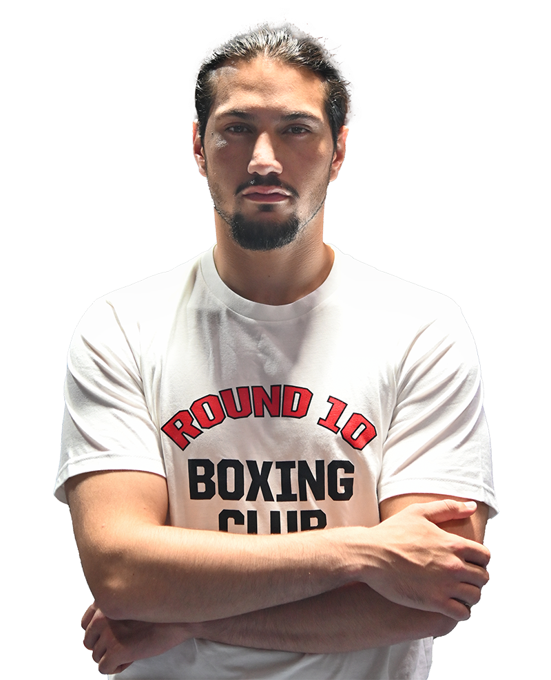 A confident man with sharp dark hair and a distinguished goatee stands with arms crossed at Round 10 Boxing Club in Al Quoz, Dubai. Clad in our signature white club t-shirt, he embodies the spirit of excellence and discipline that defines our gym—an ideal setting for those committed to mastering the art of boxing under expert guidance.