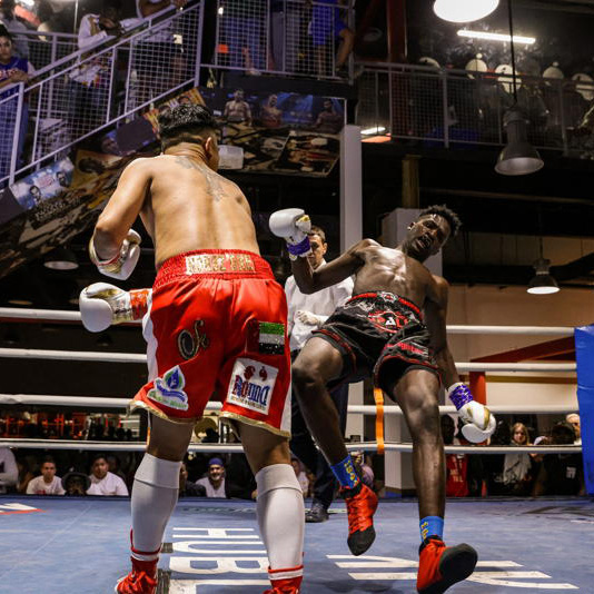 Elite fighters clash in the ring: red shorts charges, black shorts retreats. Referee and crowd watch from our Dubai boxing arena.